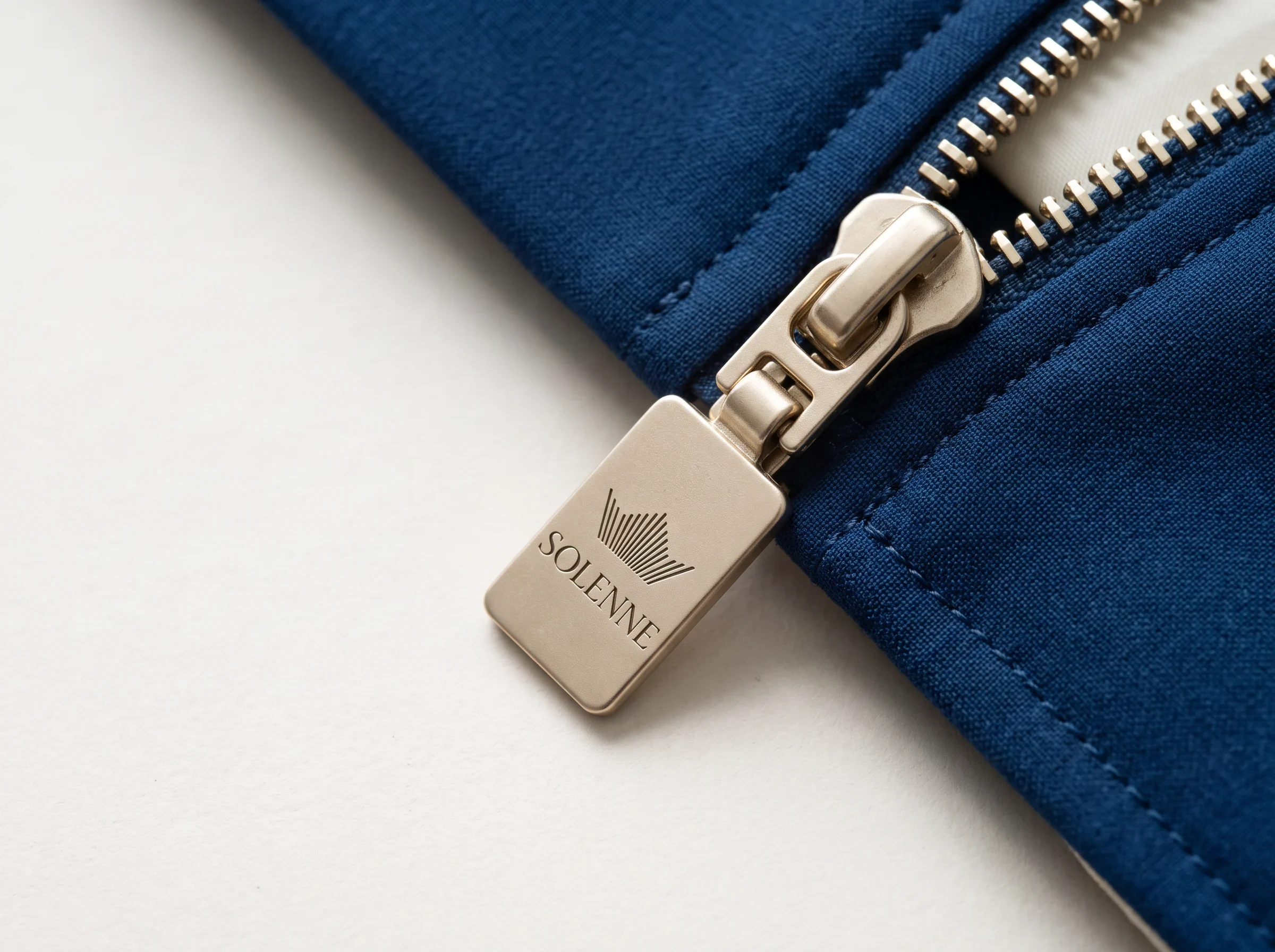 Solenne wordmark and emblem engraved on a champagne metal zipper pull.